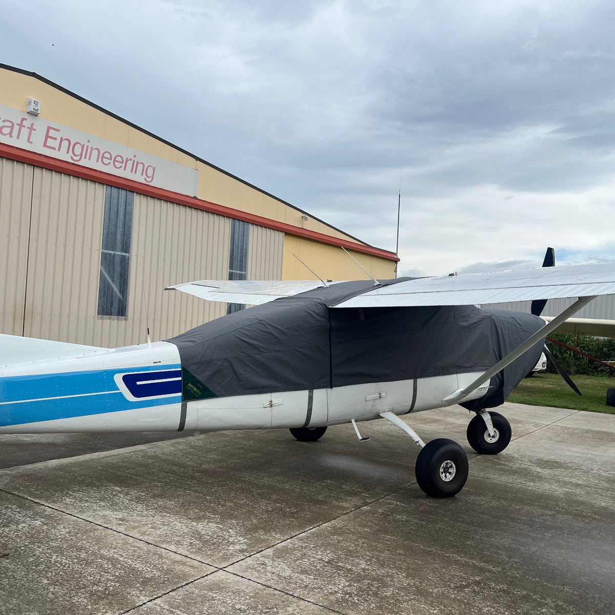 Side view of a white and blue Cessna C206 fitted with a charcoal canopy cover extending over the cockpit, nose and wing roots.