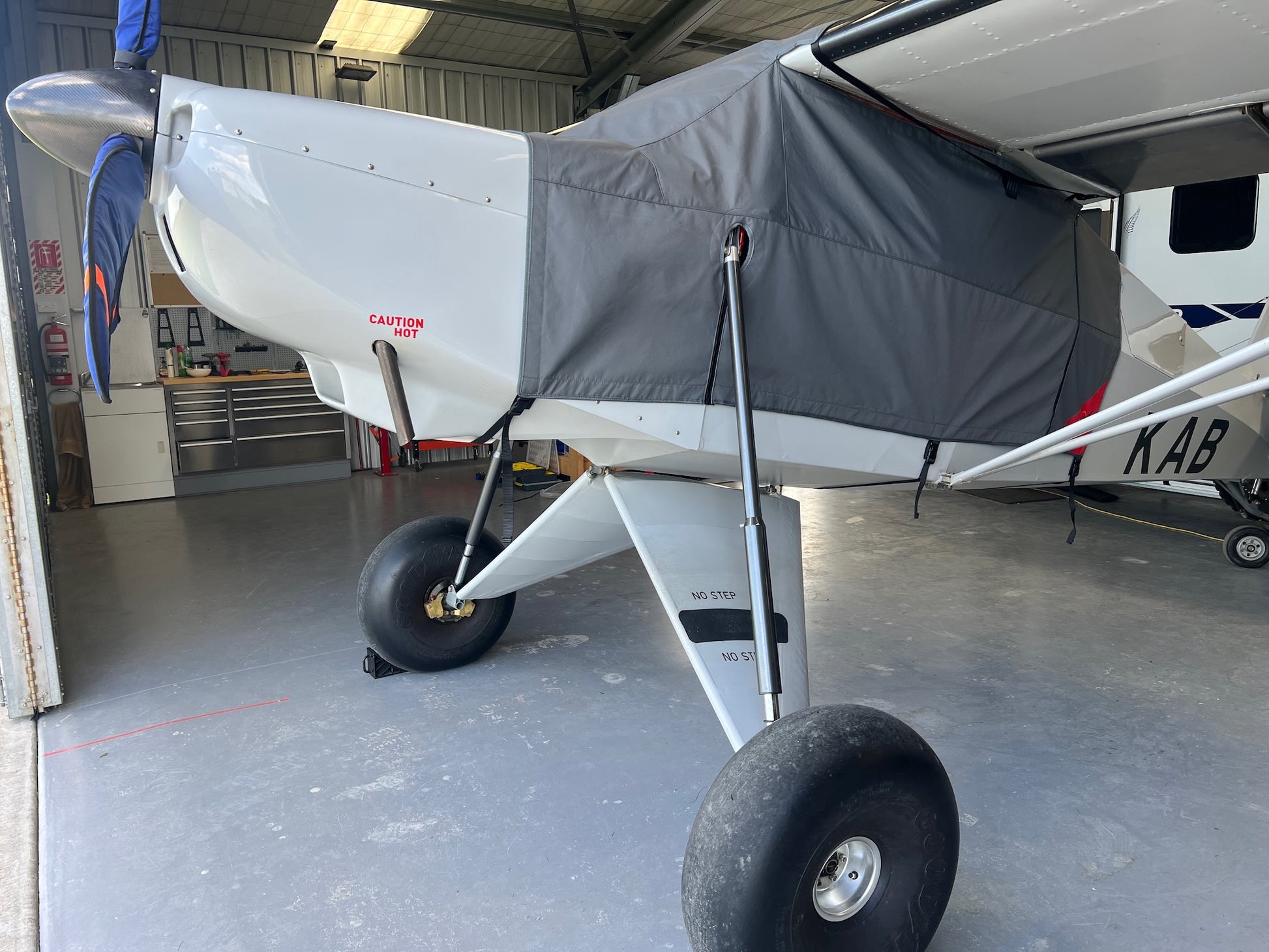 Charcoal Aerosew canopy cover fitted to a white Hornet Super STOL aircraft inside a hangar. The tailored cover protects the cockpit, windows, cabin and wing-root area while the aircraft is parked.