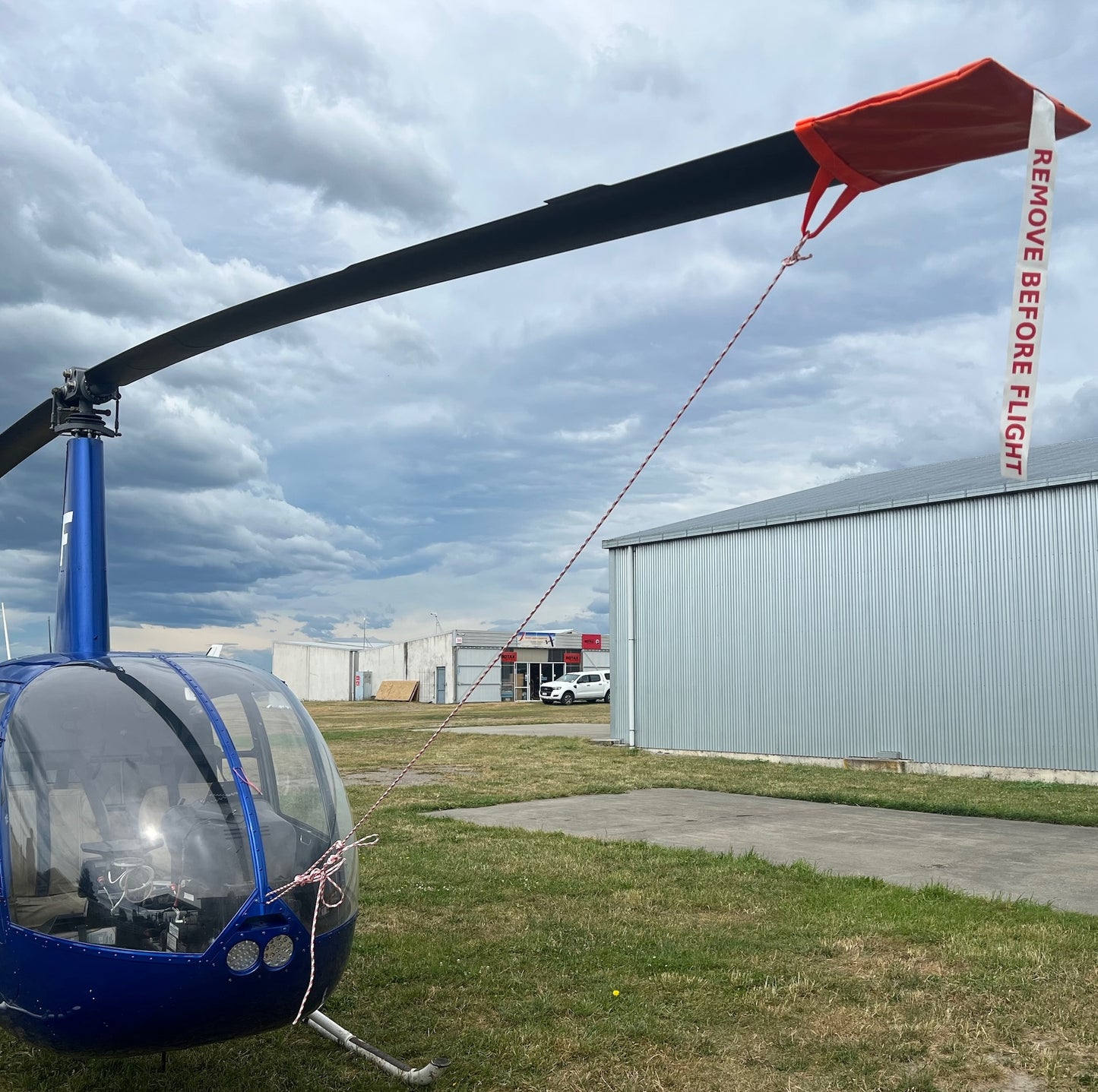 Robinson R44 Helicopter with a aerosew pvc tiedown with 'Remove Before Flight' flag on a grassy area near a building under a cloudy sky.